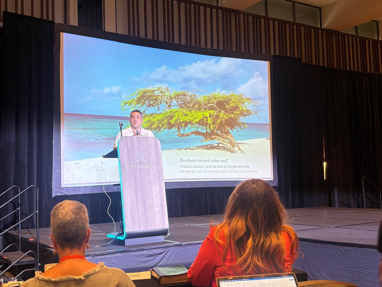 Rendell de Kort at the St. Regis podium, title slide reading Resilient toward what end? Aruba's tourism and the trap of the perfect shell