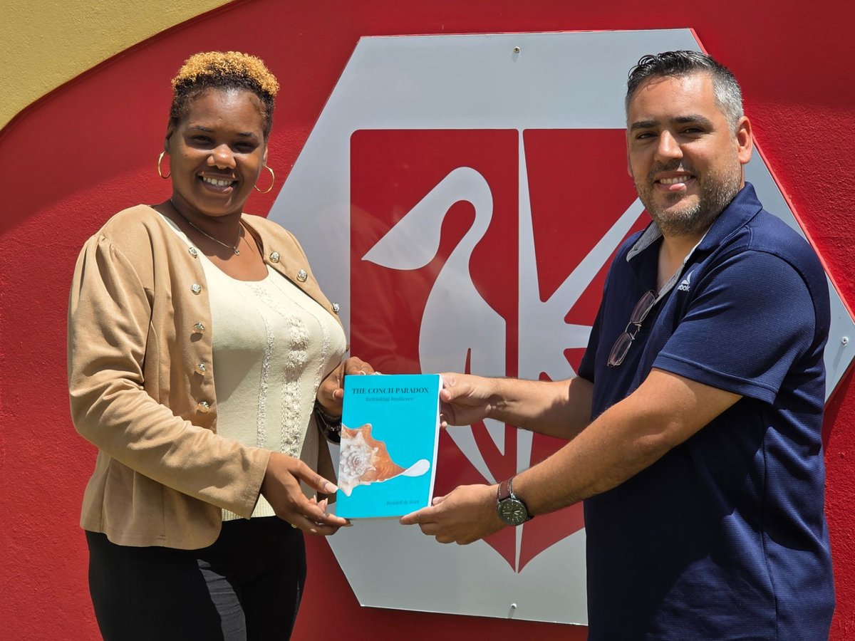 Rendell de Kort and USM Librarian Stacey Thomas holding The Conch Paradox together in front of the red and white University of St. Martin pelican logo